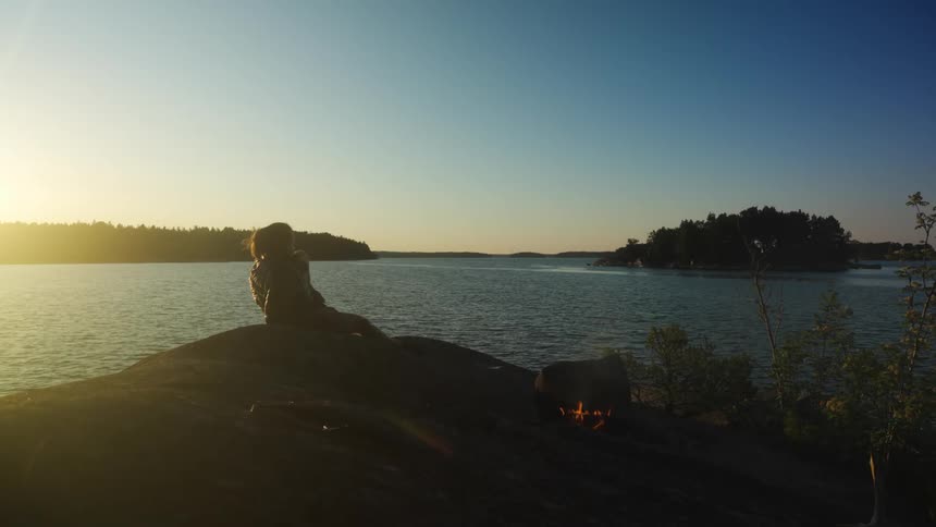 Silhouette of a woman sitting on a rock next to a campfire and on the edge of a lake with a sunset on the horizon