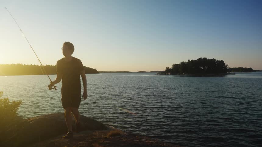 A person holding a fishing rod climbs from he shoreline of a lake at sunset
