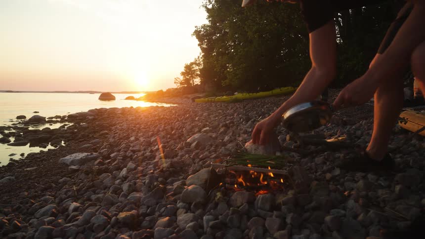 A small campfire burns on a rocky shore with the sun setting over the water in the background