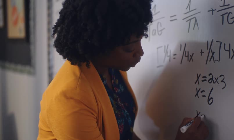 A woman with curly hair writing mathematical equations on a whiteboard while wearing a bright yellow blazer