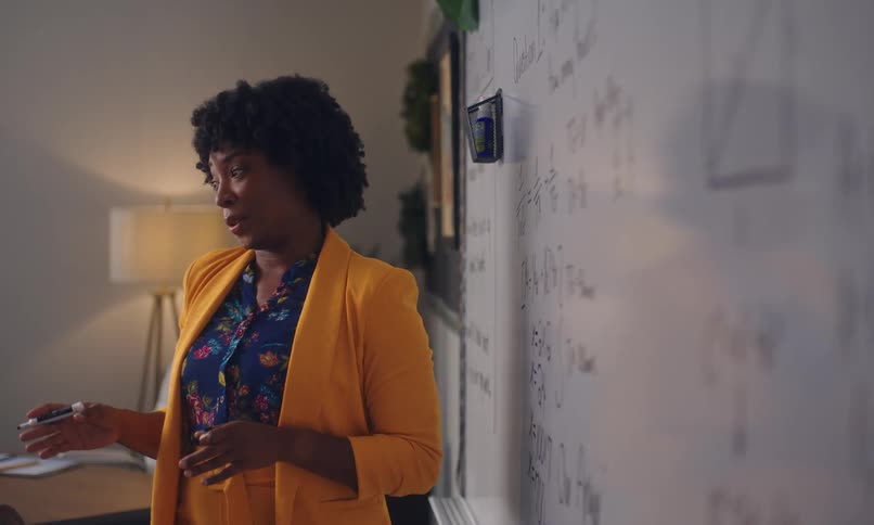 A teacher wearing a bright yellow outfit talking and writing on a whiteboard covered in mathematical equations and diagrams