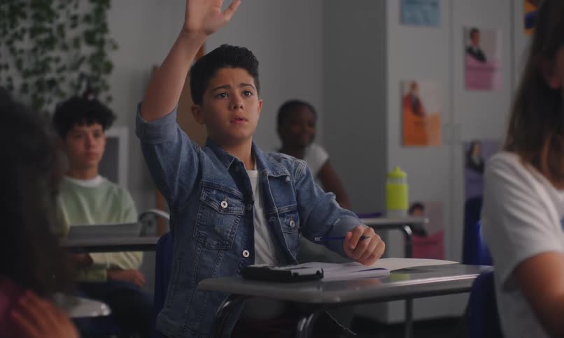 A classroom setting where a young boy is raising his hand to answer a question while other students are seated at their desks