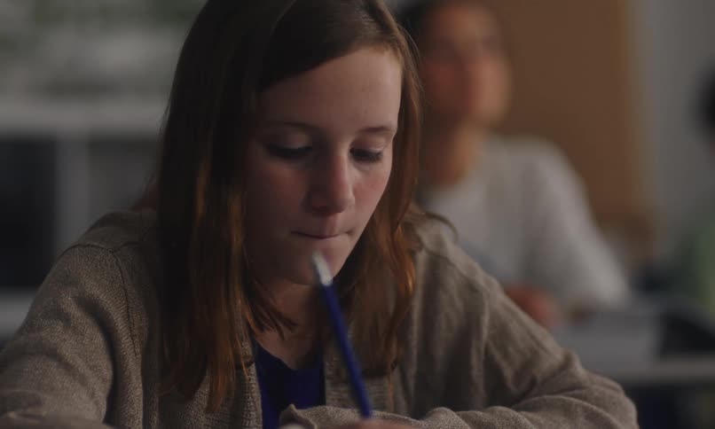 A student sitting on a classroom takes notes while listening to the teacher