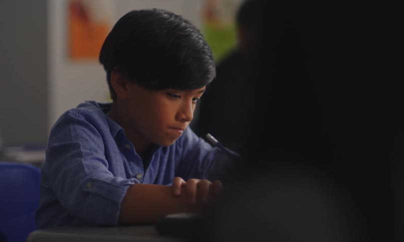 A latino boy writes on his notebook then looks up in a classroom