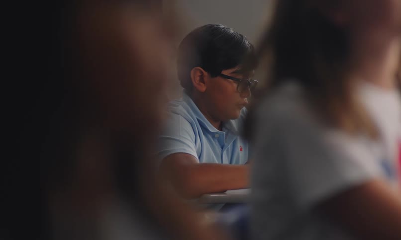 A young boy is studying in a classroom with two classmates blurred in the foreground