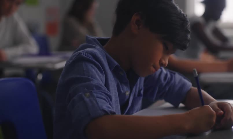 Portrait of a concentrated student writing and in the background with other students in the classroom raising their hands