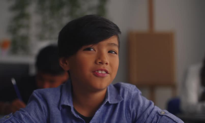 A little boy in a classroom raises his hand up and then starts talking