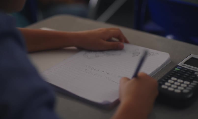 Student sitting at a desk writing with a pencil in a notebook with a calculator next to it