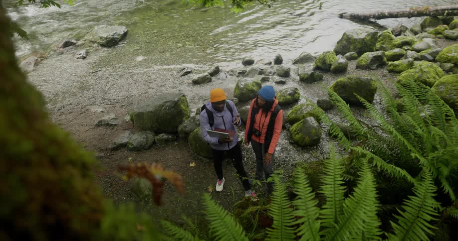 A couple holding a laptop stand by a riverbank surrounded by rocks covered in moss