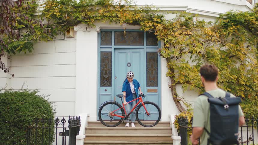 Two friends salute each other while one of them carries a bicycle out of a house