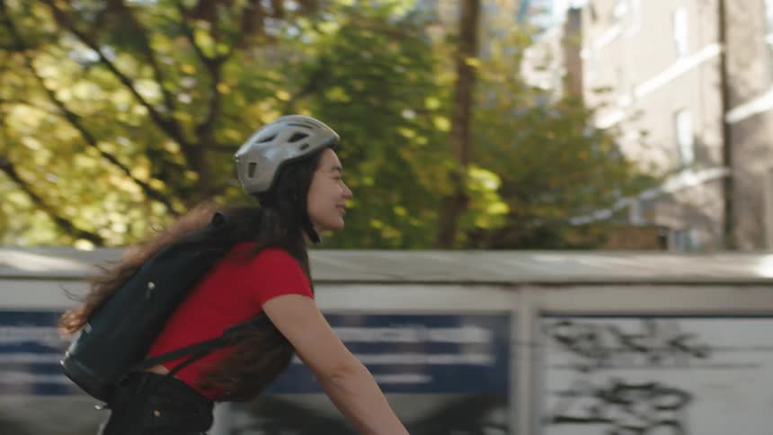 A woman with long hair riding a bicycle in a city wearing a helmet and a red shirt with a backpack on her back