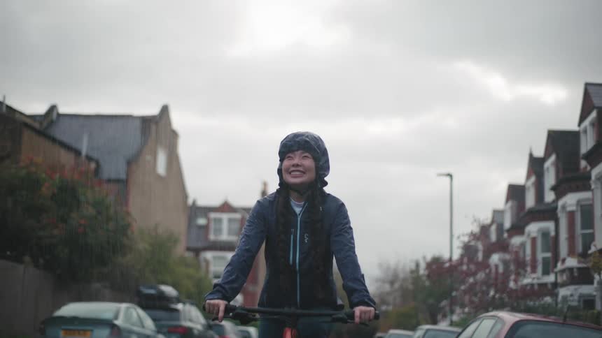 An asian woman rides her bicycle down a suburban street in the rain