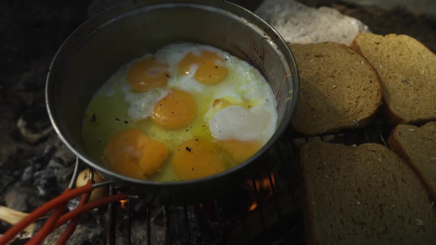 Cracked eggs in a frying pan cooking over an open flame beside rye bread on an outdoor camping grill