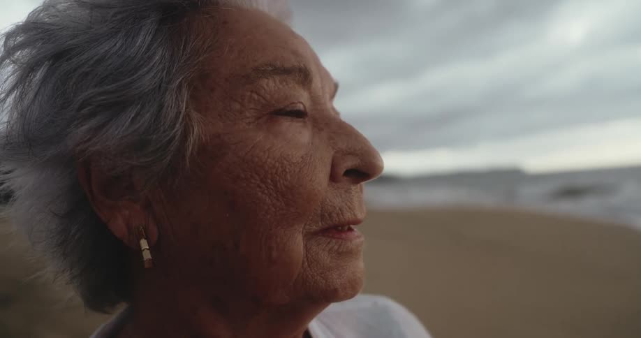 A woman with gray hair and an earring is standing by the beach with the ocean in the background
