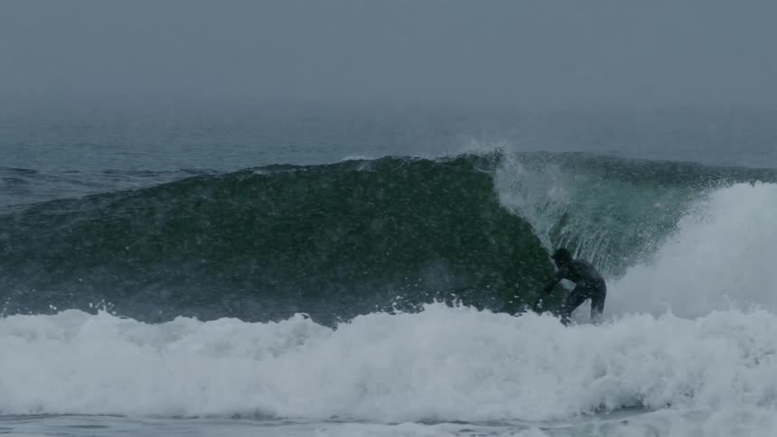 A surfer rides a wave in the ocean water spraying around under a snowstorm