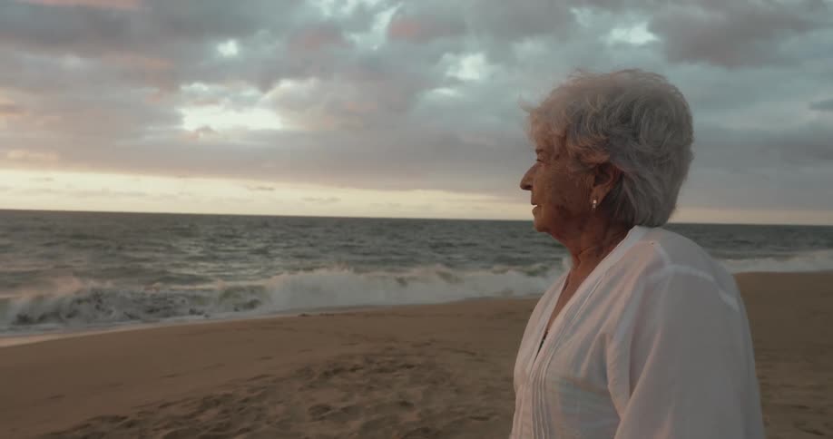 An elderly woman with grey hair stands on a beach gazing out at the ocean where gentle waves crash onto the shore