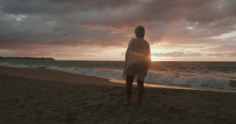 A woman stands on the beach and stares off into the sunset at the horizon