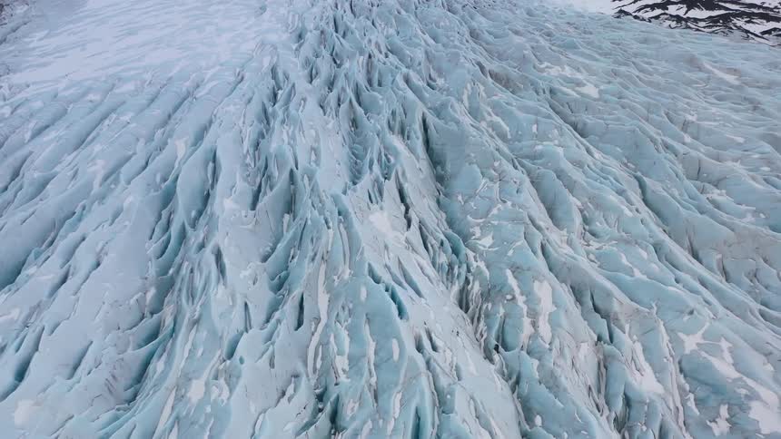 An aerial view of a glacier with deep blue crevasses between the ice surrounded by snow covered mountains