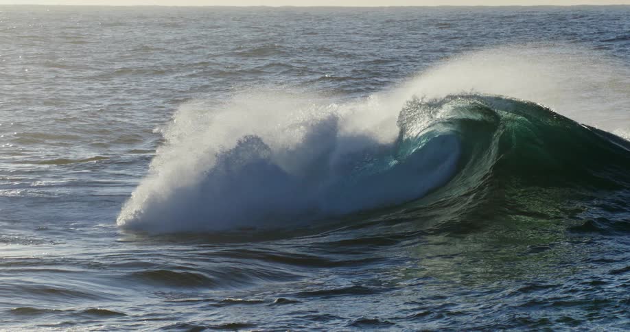 Large wave flowing across the ocean surface breaks creating a spray of white water
