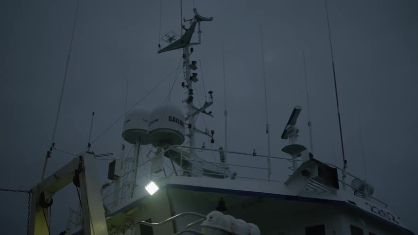 A ships upper structure at twilight with navigation equipment and antennas against a cloudy sky illuminated by a single bright light on the deck
