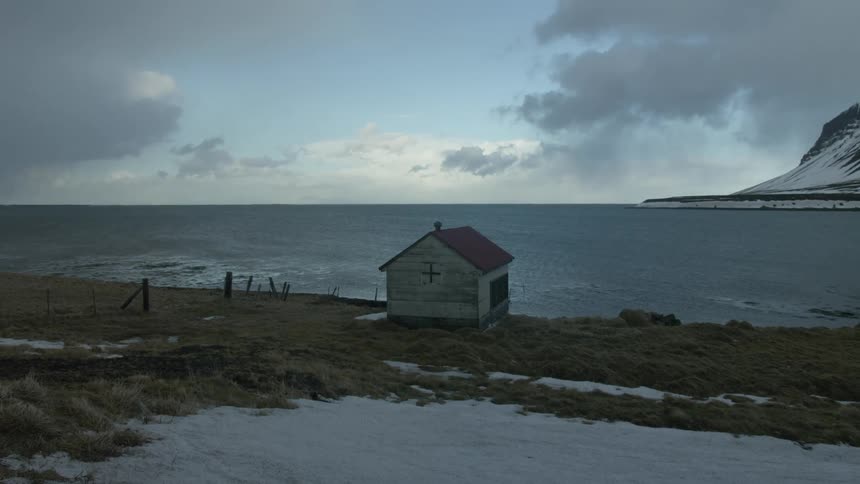 Lonely house is situated on a grassy snowy shore with a mountain in the background and a cloudy sky above