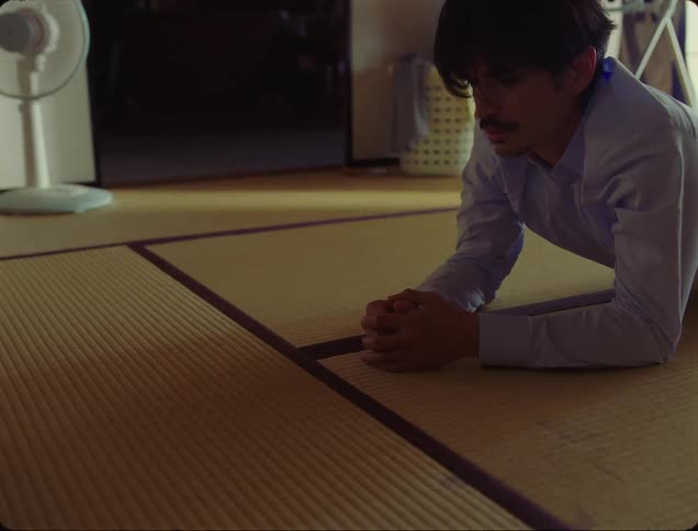 A child in blue socks walks past an adult lying face down on a tatami mat floor the man stands up and follows the child