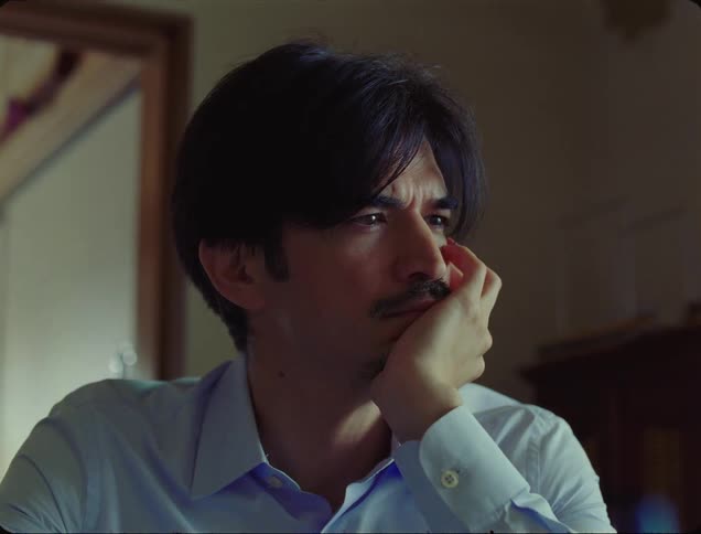 A man sits in a house and stares into space before turning his head to look behind him
