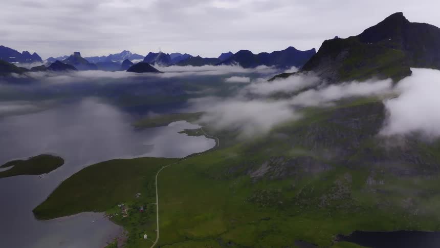 A serene lake nestled among green fields flanked by rugged mountains and partially obscured by lowhanging clouds