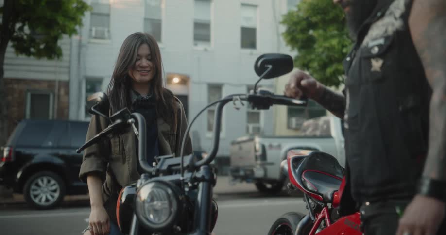 A woman sitting on a black motorcycle talking to a tattooed person standing beside it