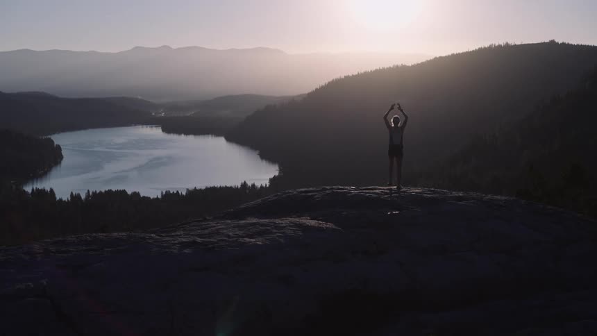 Silhouette of a person on a mountain peak with arms outstretched upwards against a backdrop of a lake and sunrise on the horizon