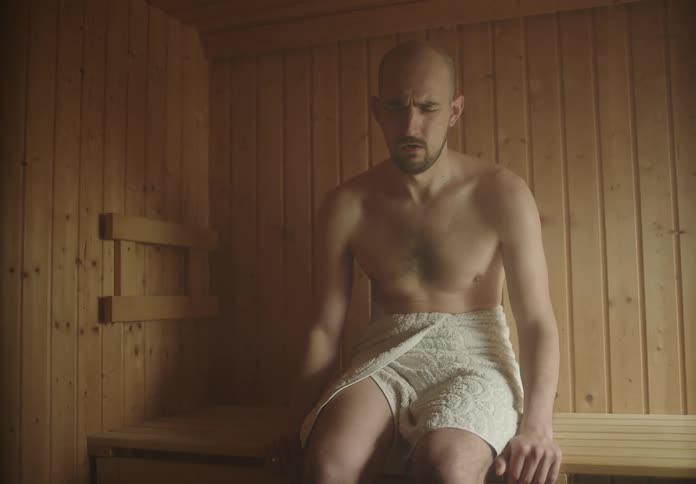 A man taking deep breaths in a sauna while looking anxious is joined by another