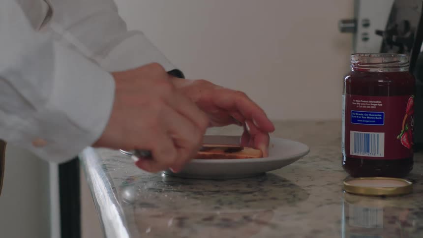 A closeup of a hand holding a knife spreading jam on a slice of bread that is on a plate