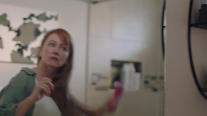 A woman brushes her red hair while looking in a bathroom mirror