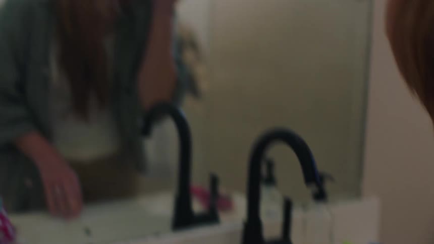 A woman reflected in a bathroom mirror applying makeup to her face with a brush