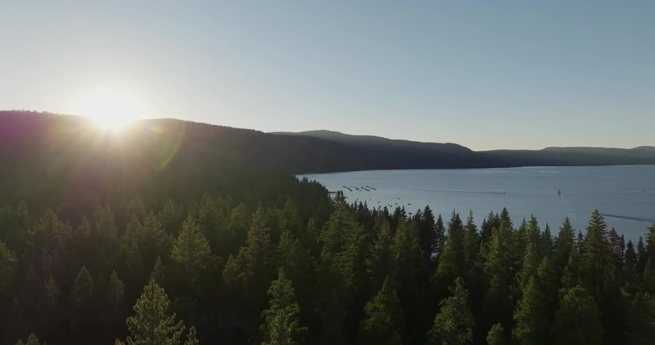 Aerial landscape of a lake surrounded by mountains and forests with conifers and the sun on the horizon