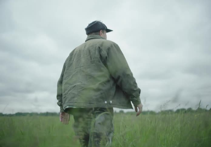 A man wearing a jacket and a hat walking on a field of green tall grass