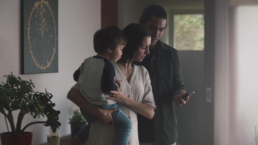Mother is holding child at home when father joins them and shows her a smartphone