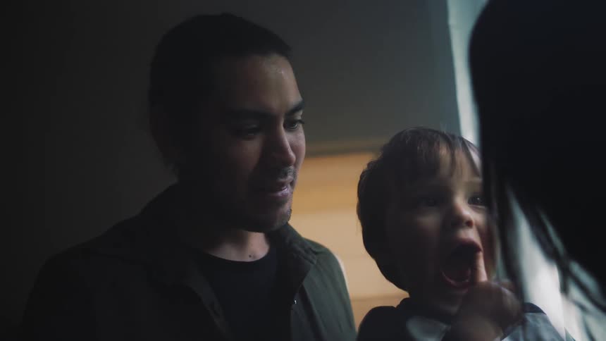 A toddler boy putting his finger in his mouth then smiling at the person in front of him with his father behind him
