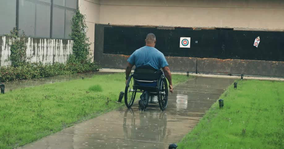 A black man in a wheelchair rolls over a sidewalk as rain falls