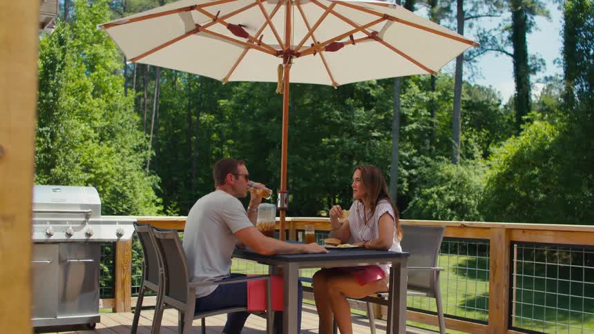 Man and woman sitting on a wooden deck and at a table with an umbrella talking and drinking