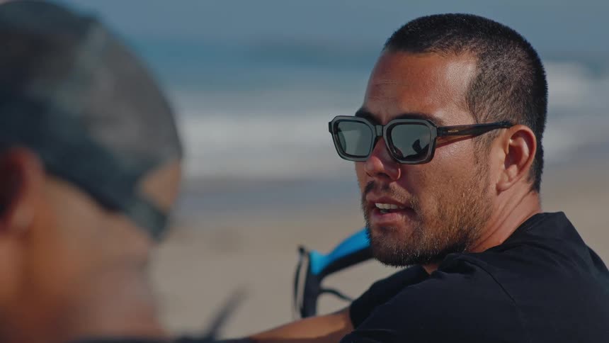 Man sitting with sunglasses talking to a person there while on the beach