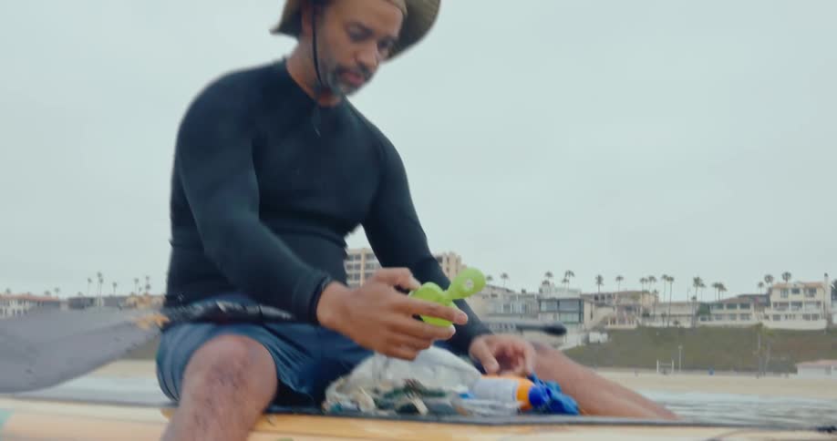 Man sitting on a surfboard collecting trash and plastic in the ocean waters