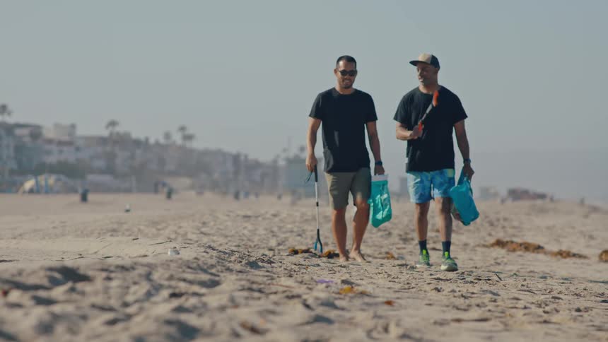 Two men talk while picking up litter on the beach near a coastal town