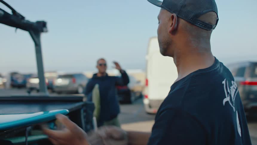 A man places a surfboard on the back of a truck and then shakes hands with a friend