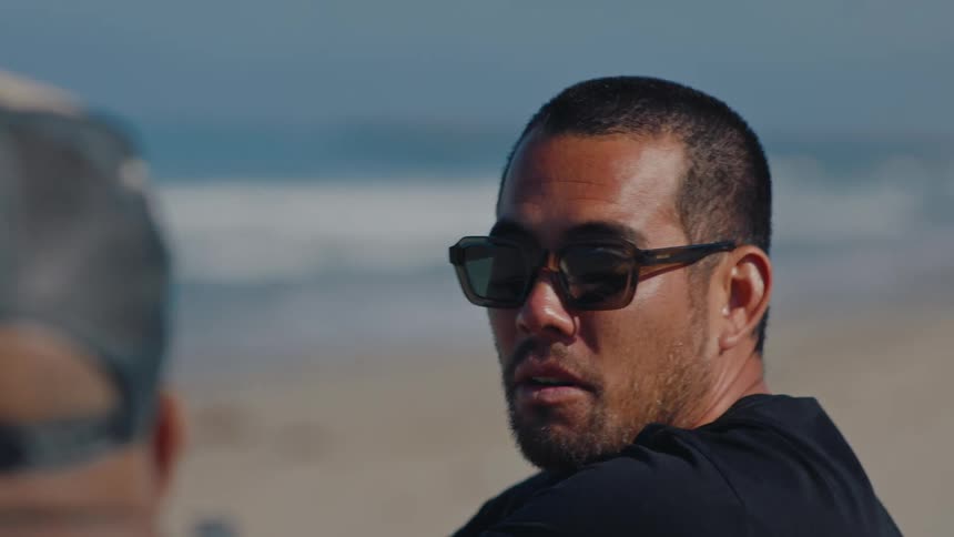 A mans face wearing sunglasses and he is on a beach talking to someone in front of him