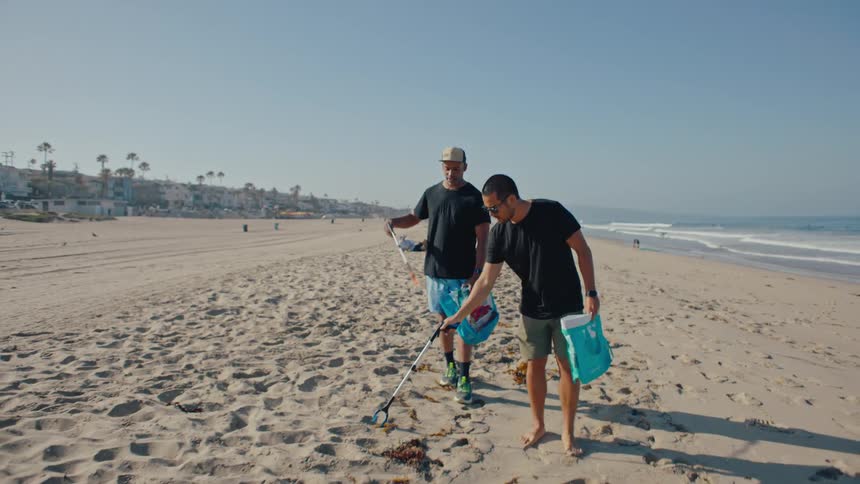 Two young men walk along a beach and pick up trash