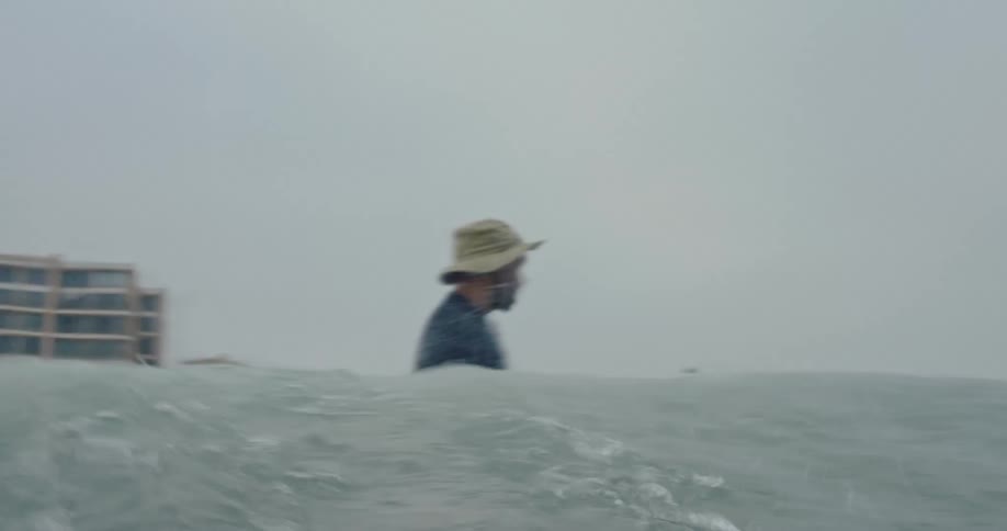 A man in a hat is surfing a small wave and then he falls and on the shore there are several large buildings