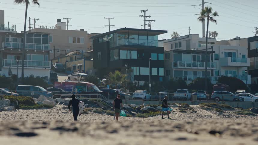 A group of people carrying surfboards walking on a beach next to residential buildings on a busy street