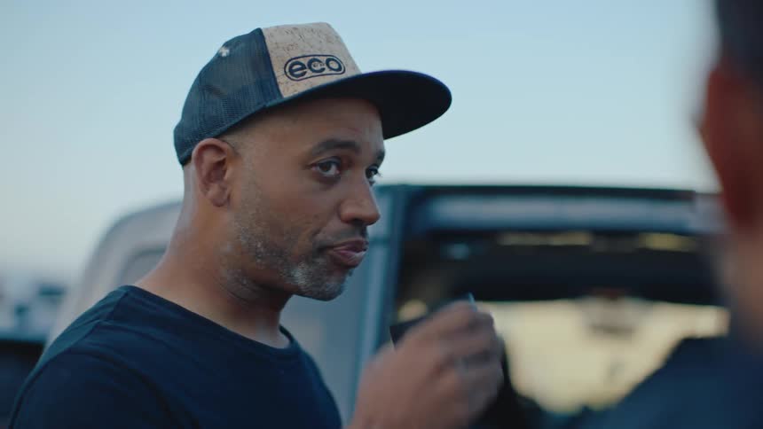 A man wearing a cap who is drinking something and then talking to someone in front of him