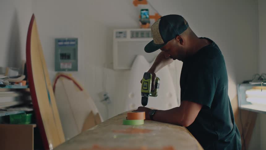 Young man repairs a surfboard using a drill and colorful tape